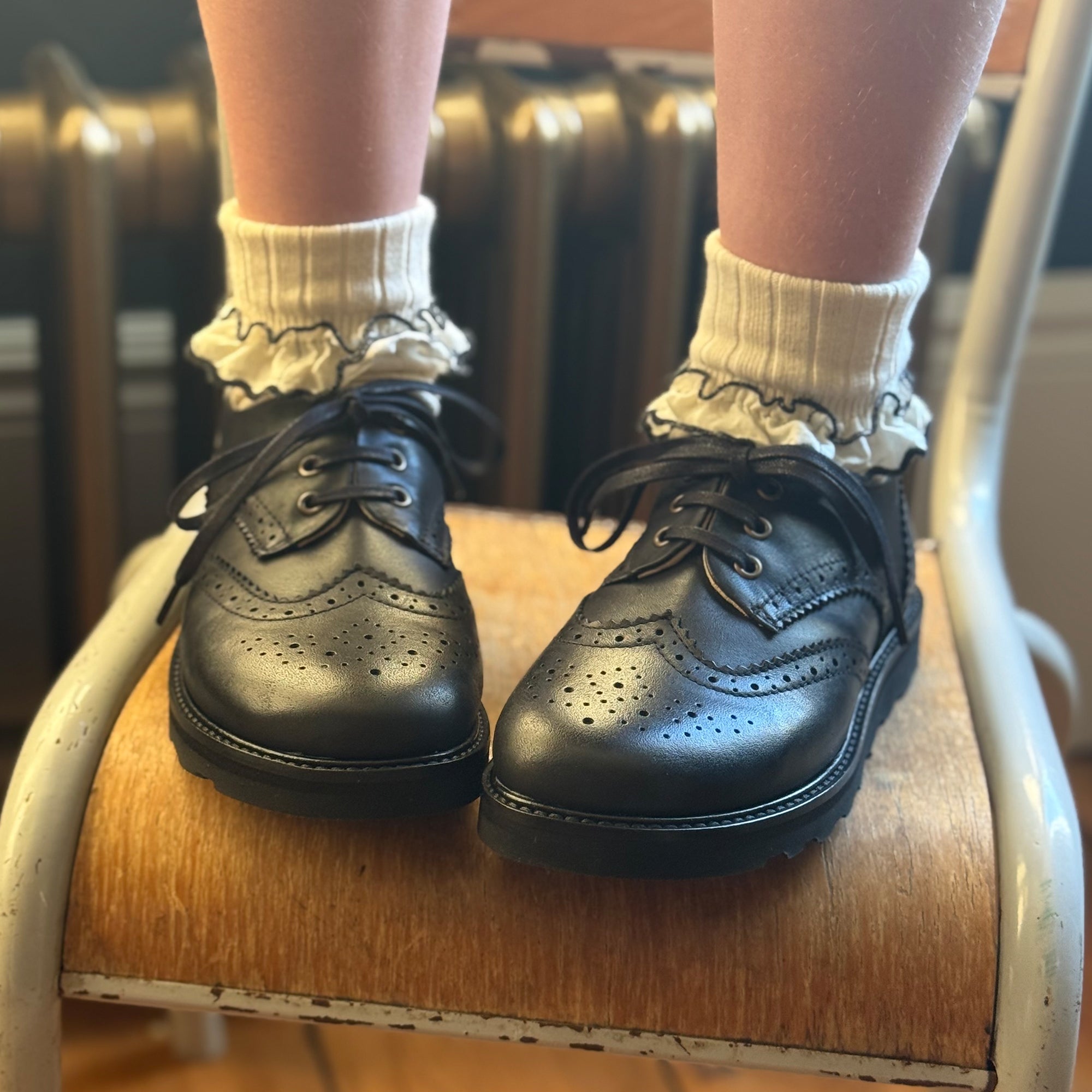 Black Young Soles Brando back to school shoes with ruffled white socks on a wooden chair.