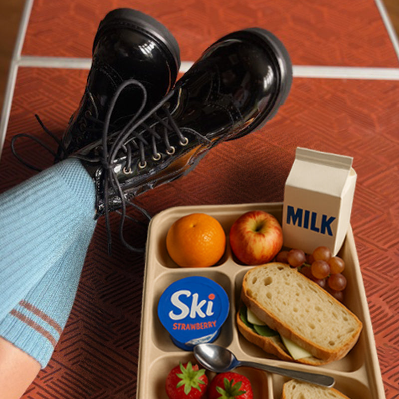Person wearing black patent Young Soles Sidney brogue boots and light blue socks with a lunchbox containing a sandwich, fruits, and a carton of milk on a red tiled table.